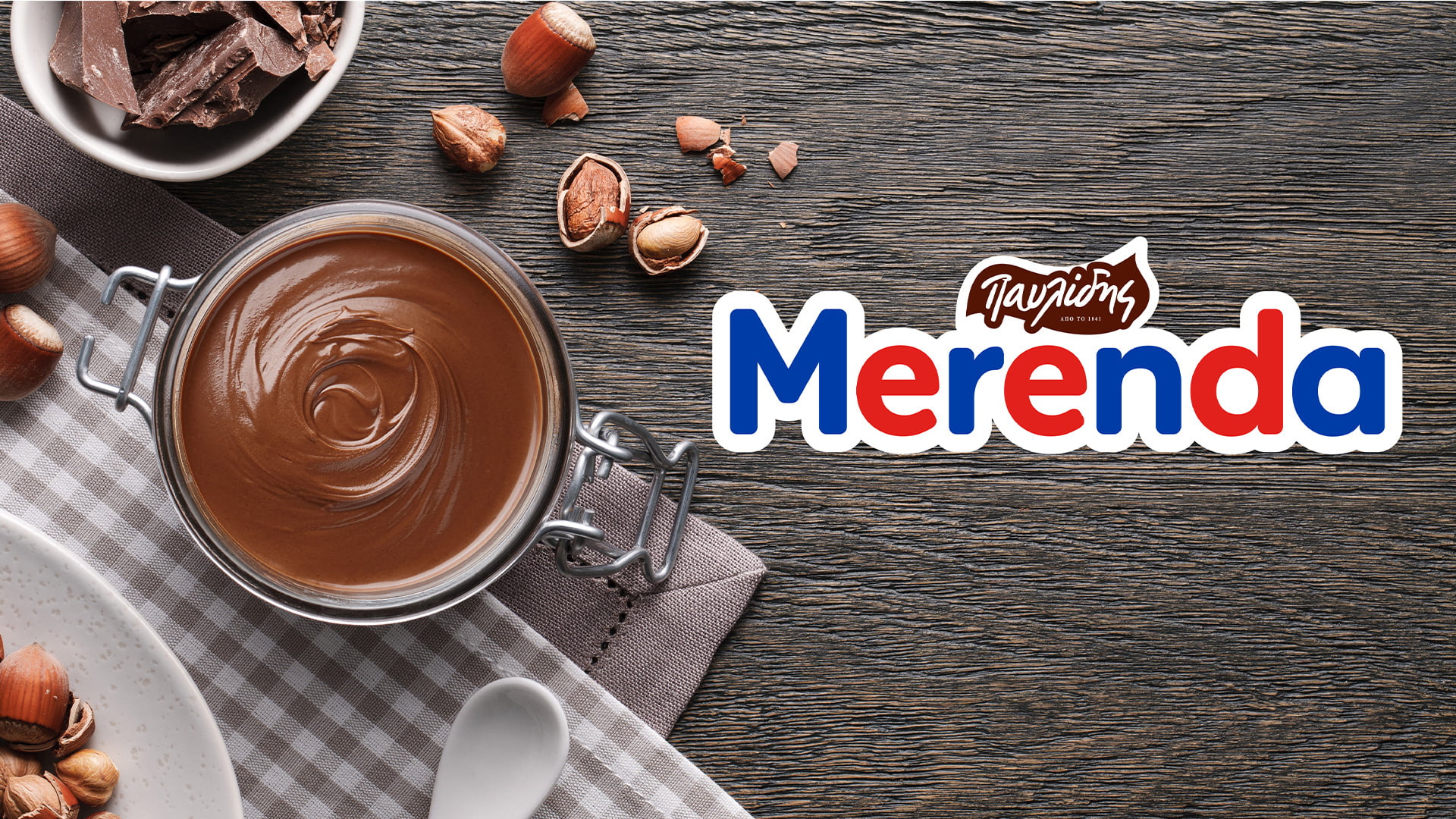 The redesigned Merenda (by Pavlidis) logo on a dark wooden table, next to an open jar of chocolate-hazelnut spread, surrounded by hazelnuts and chocolate chunks.