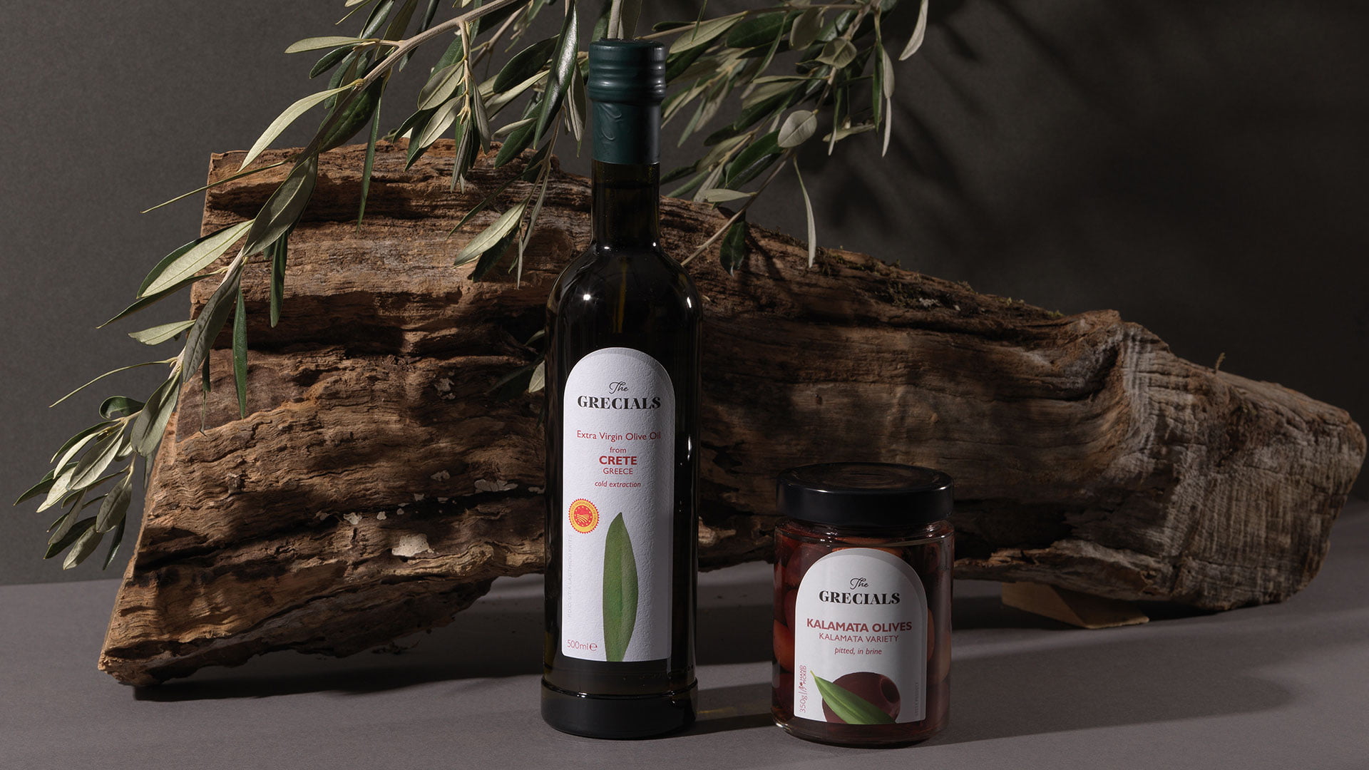 A product shot of The Grecíals, showing a bottle of Extra Virgin Olive Oil from Crete and a jar of Kalamata Olives, staged with rustic driftwood and olive branches.