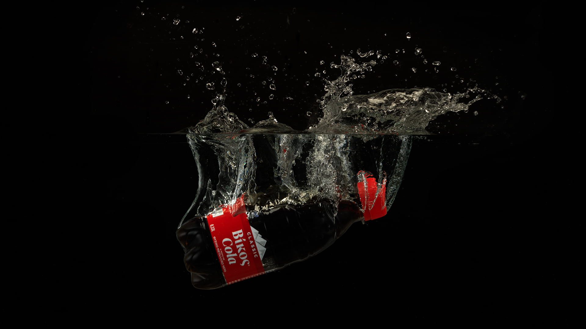 A high-speed photograph of a Classic Vikos Cola PET bottle splashing sideways into dark water, creating a large, dynamic splash against a solid black background.