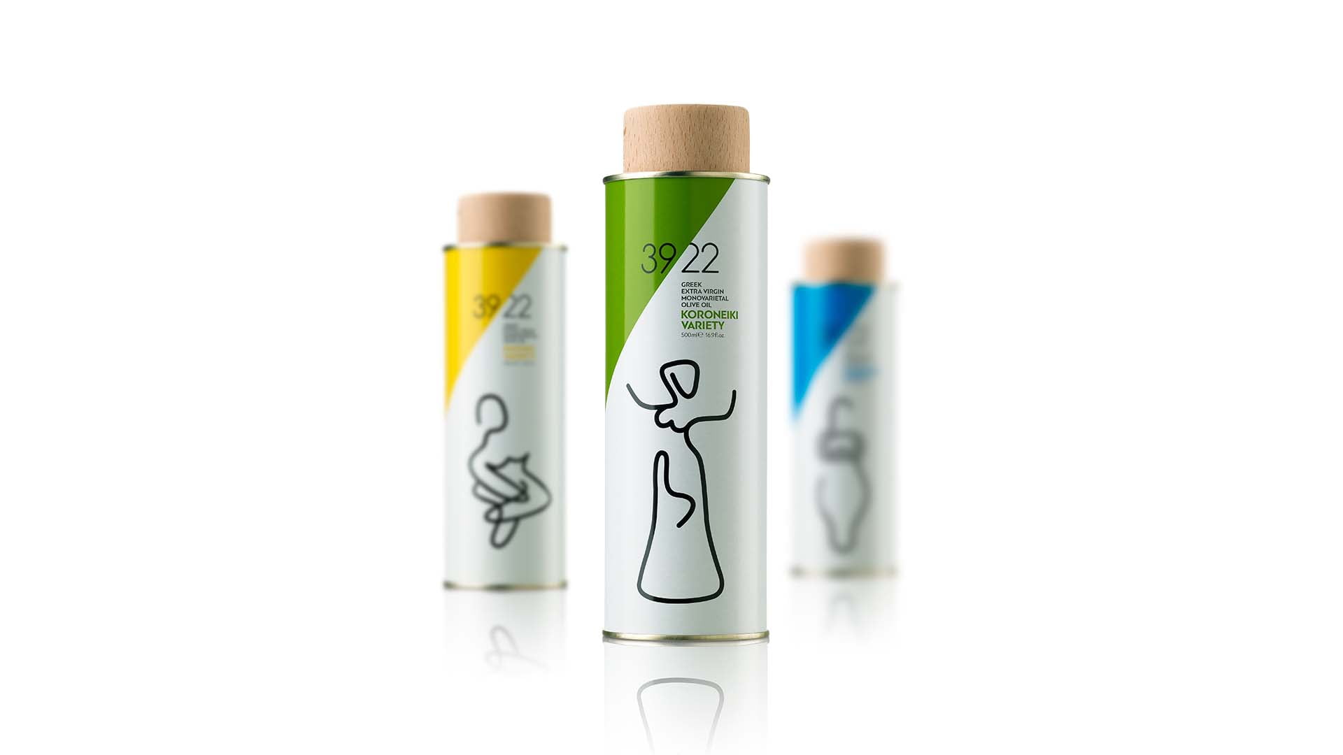 A product lineup of 39/22 Greek Extra Virgin Olive Oil tin bottles, with the green "Koroneiki Variety" in sharp focus in the center, and yellow and blue versions blurred in the background, all on a white studio surface.