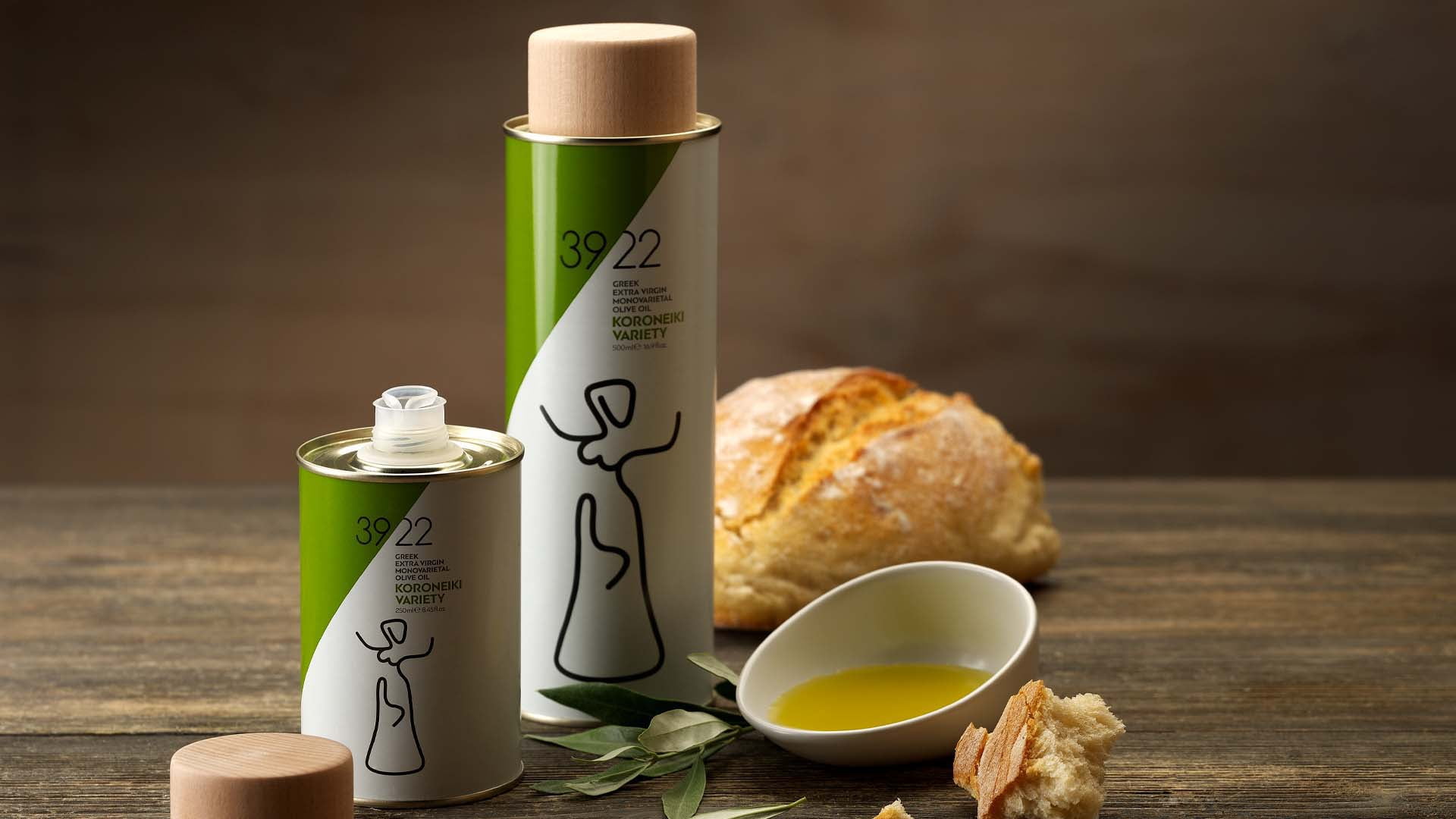 Two 39/22 Koroneiki Variety olive oil tin bottles, in large and small sizes, styled on a rustic wooden table with fresh bread, olive leaves, and a white bowl of dipping oil.