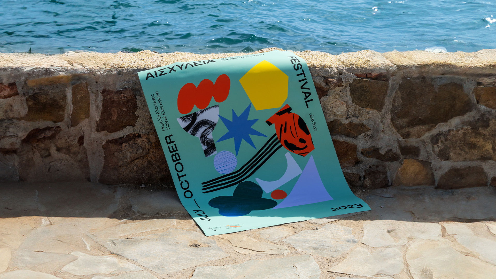 A mint green poster for the Aeschylia Festival 2023, featuring an abstract collage of colorful shapes and graphics, lying on a stone path next to a low stone wall with the sea in the background.