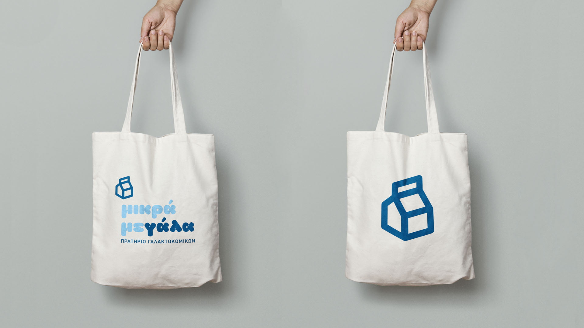 A mockup of two white canvas tote bags for the "μικρά μεγάλα" (mikra megala) dairy store: one featuring the full logo and text, and the other featuring just the large, dark blue milk carton icon.