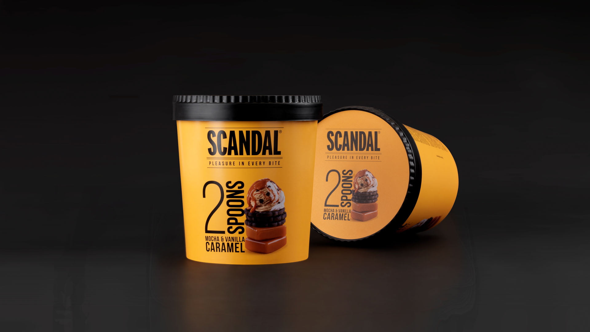 A product shot of two Scandal '2 Spoons' ice cream tubs in 'Mocha & Vanilla Caramel' flavor, showing the orange and black packaging on a reflective black background.