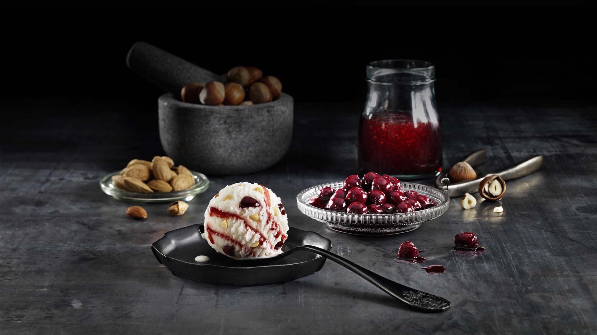 A dark and moody food styling shot for Scandal 'Vanilla Parfait' ice cream, showing a scoop of vanilla ice cream with cherry drizzle, surrounded by ingredients like preserved cherries, almonds, and hazelnuts.