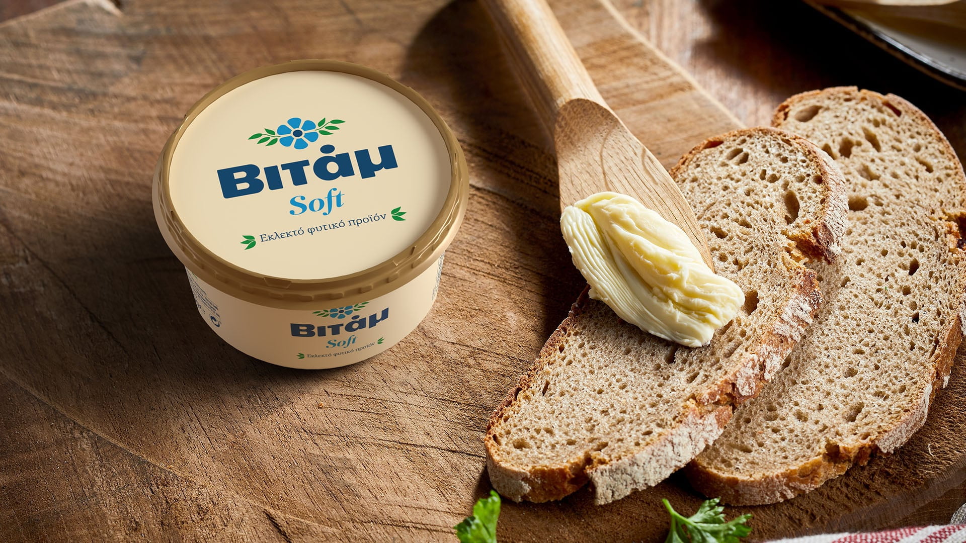 A tub of the rebranded Vitam Soft plant-based spread on a wooden cutting board, shown next to three slices of rustic bread with a curl of the spread on a wooden spoon.